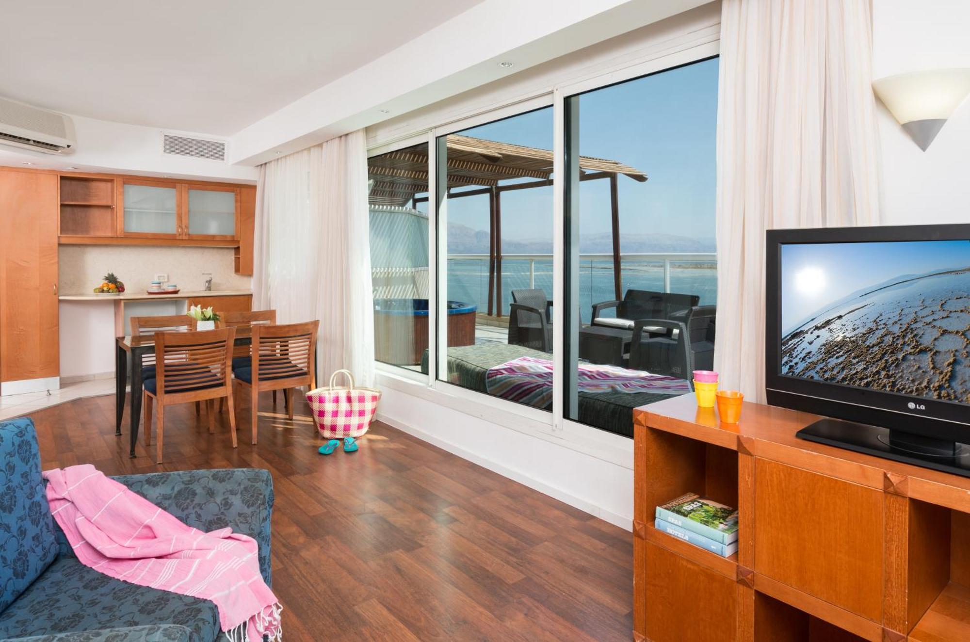 Leonardo Club Hotel Dead Sea Neve Zohar Extérieur photo The photo depicts a modern living space with a view. On one side, there is a comfortable seating area with a couch and a television cabinet. The TV is turned on, showing a scenic image. 

In the background, there is a dining area with a wooden table 